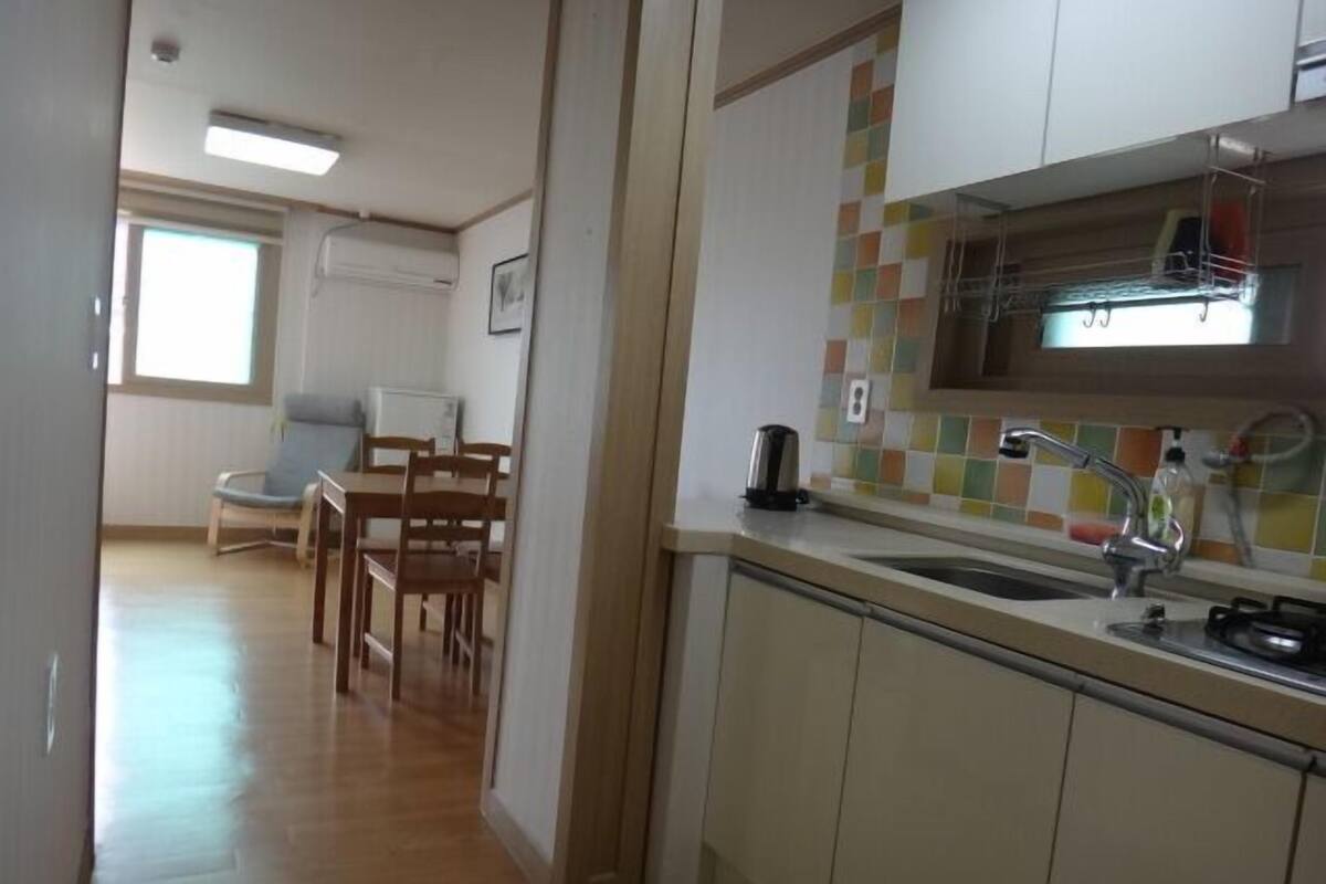 Una cocina compacta con salpicadero de azulejos, un lavamanos y una estufa. Se observa un comedor con una mesa y sillas al fondo.