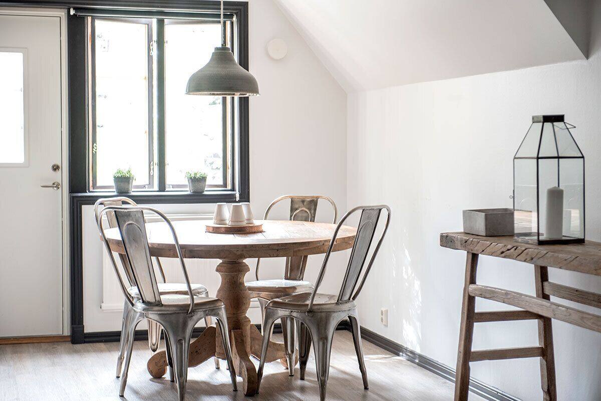 A dining area with a round wooden table, four metal chairs, a hanging pendant light, and a small side table with a lantern.