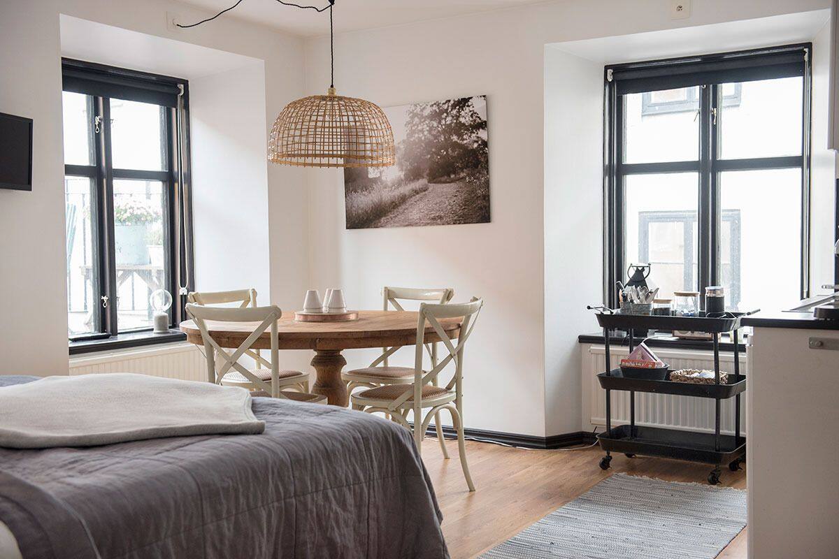 A cozy room with a bed, a dining table with chairs, a pendant light, and a black cart with kitchen items.