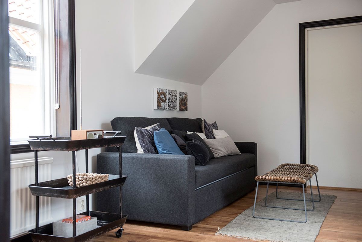 Een moderne woonkamer met een grijze bank, een klein rieten krukje, een metalen schapkas en een lijstwerk met kunst aan de muur.