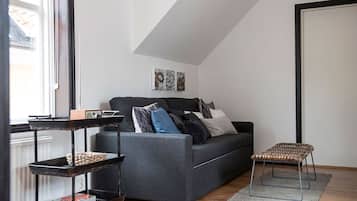 Een moderne woonkamer met een grijze bank, een klein rieten krukje, een metalen schapkas en een lijstwerk met kunst aan de muur.