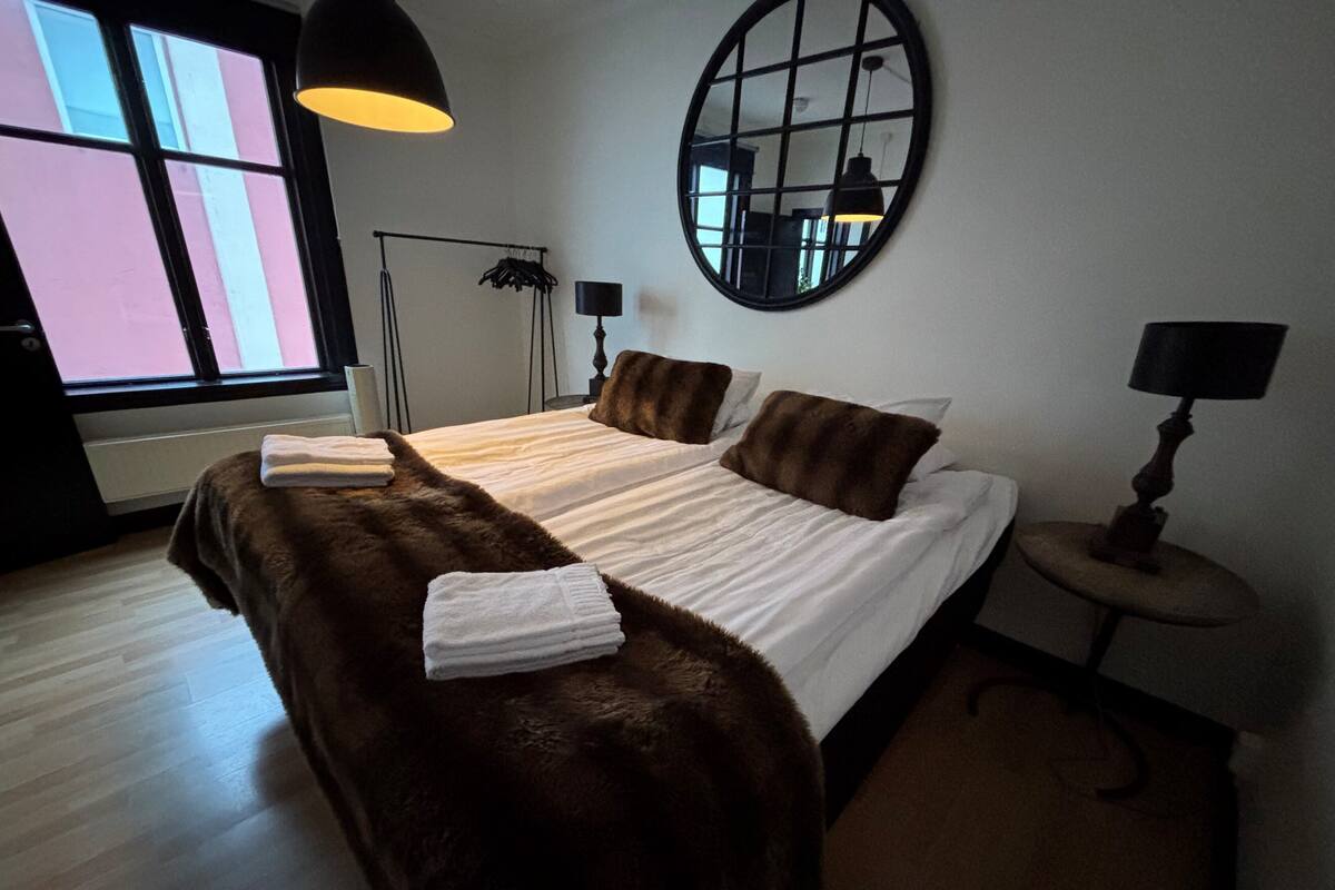 A bedroom with a large bed, a round mirror, a floor lamp, and a window with blinds.