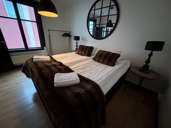A bedroom with a large bed, a round mirror, a floor lamp, and a window with blinds.