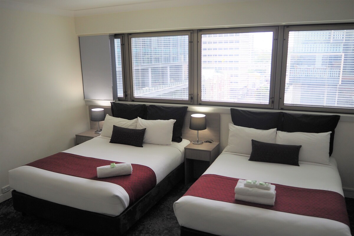 A hotel room with two beds, each with a red and white bedspread, black and white pillows, and a nightstand with a lamp.