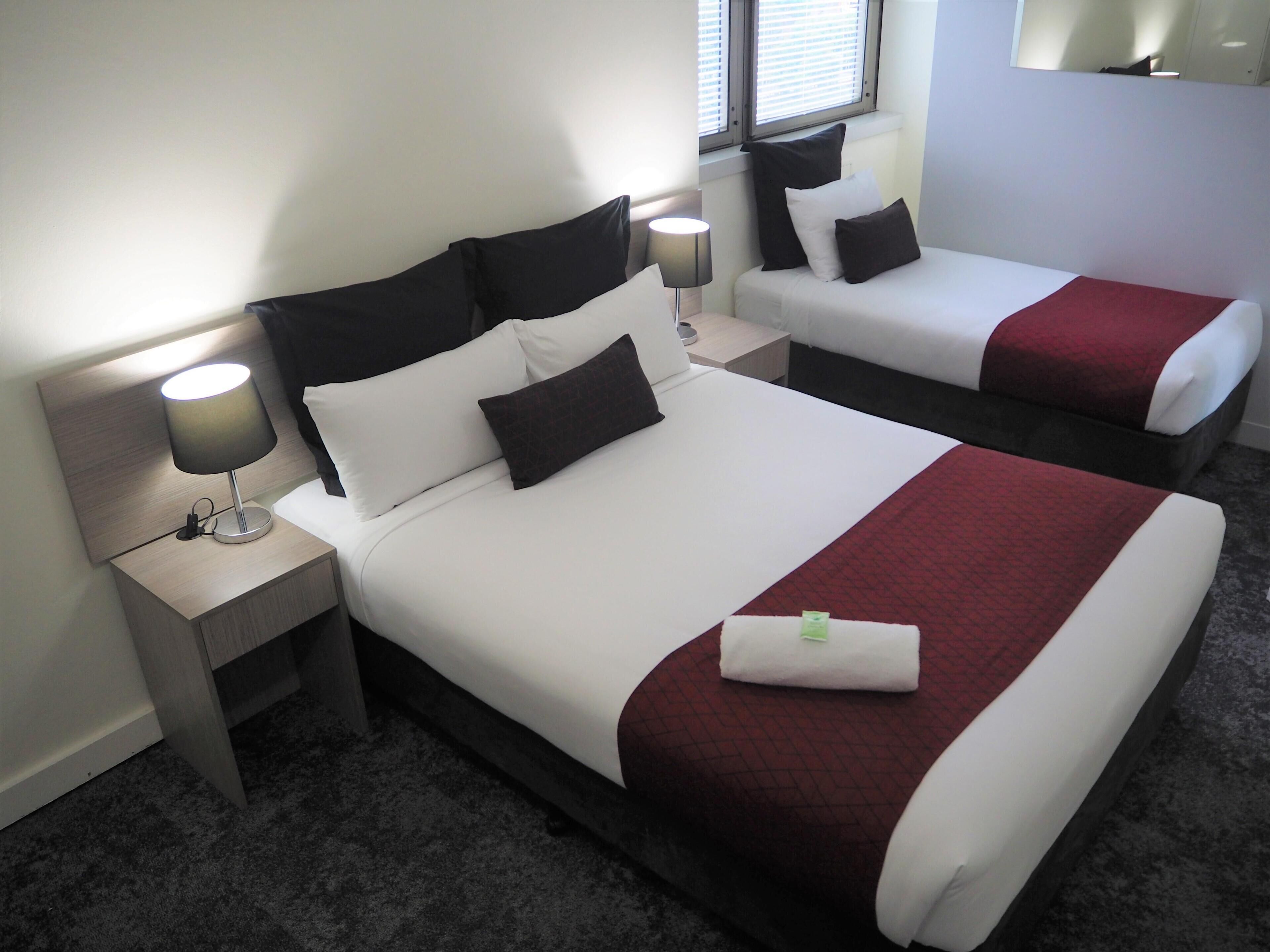 A hotel room with two beds, each with white bedding and dark headboards. There is a nightstand with a lamp on the left side of the room.