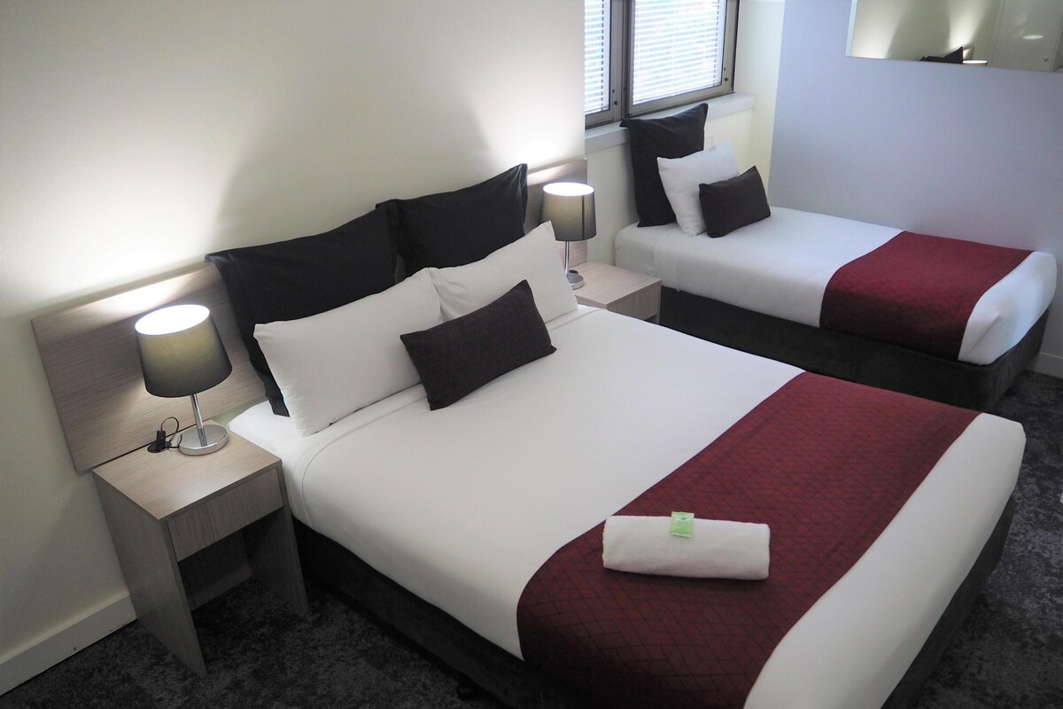 A hotel room with two beds, each with white bedding and dark headboards. There is a nightstand with a lamp on the left side of the room.