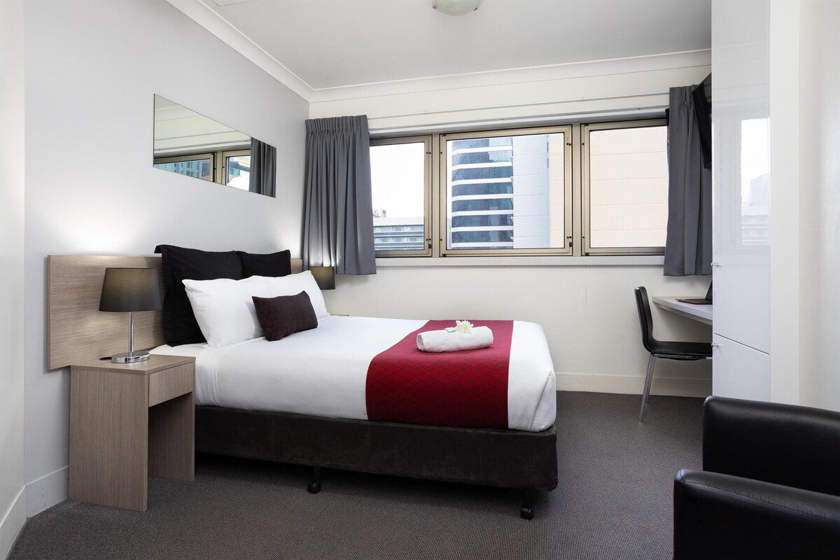 A hotel room with a bed, a desk, a chair, a lamp, and a window with curtains.