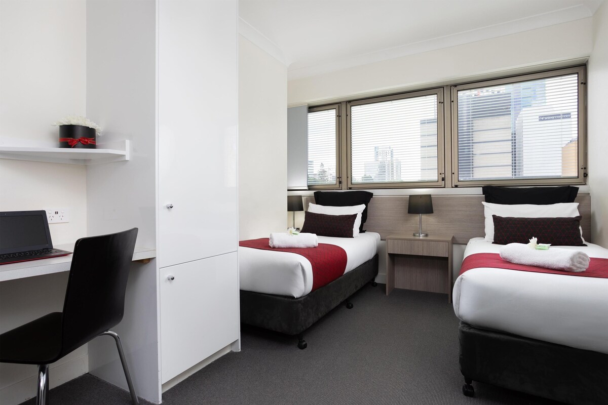 A hotel room with two beds, a desk with a laptop, a chair, and a window with blinds.