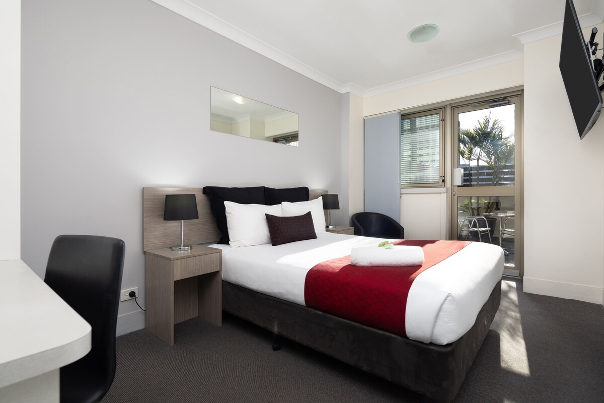 A hotel room with a bed, a desk, a chair, a TV, and a window with a view of palm trees.