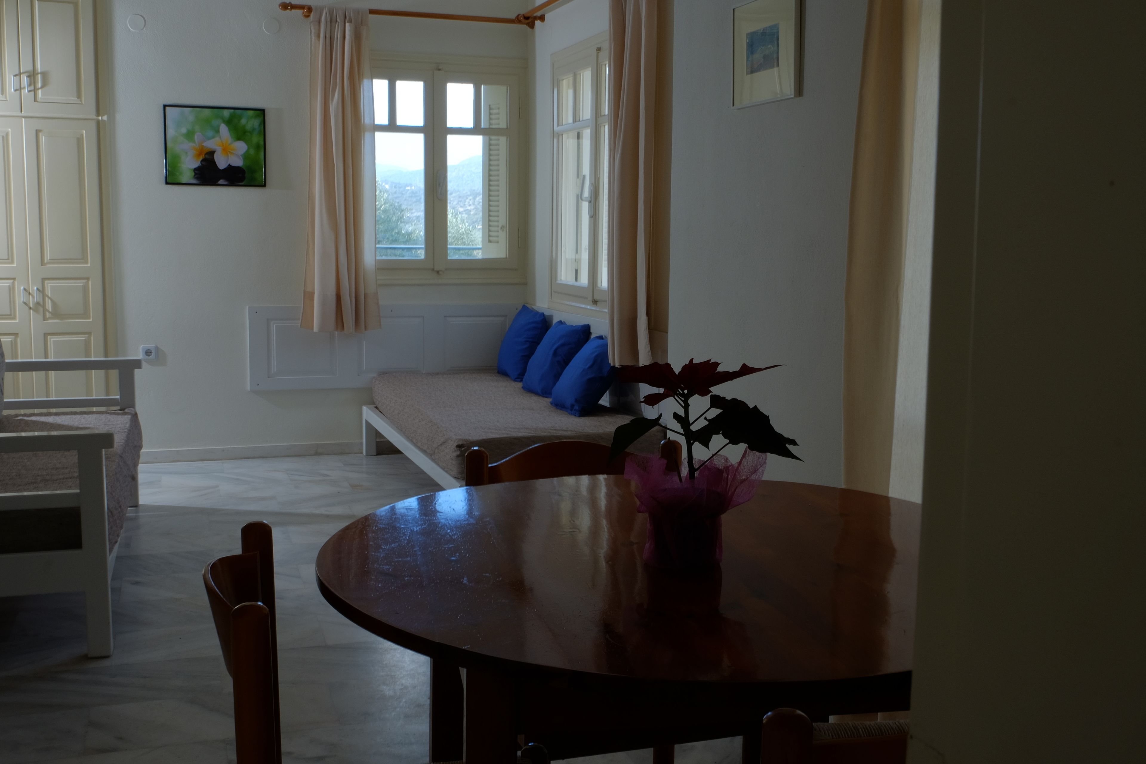 Een eetgedeelte met een ronde tafel, stoelen, een bank met blauwe kussens, een raam met gordijnen en een schilderij aan de muur.