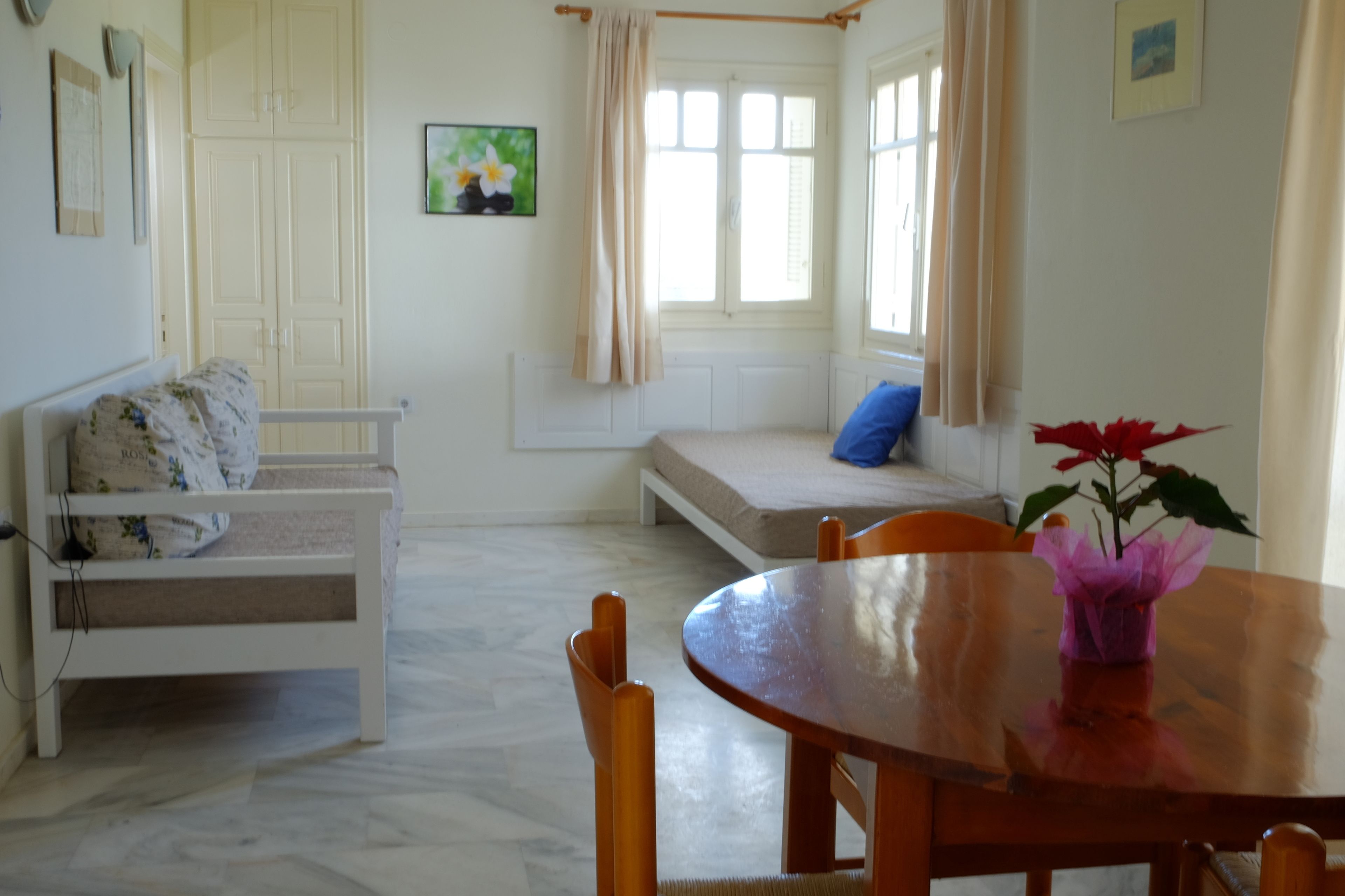A room with a wooden dining table, a white sofa, a window with curtains, and a small plant on the table.