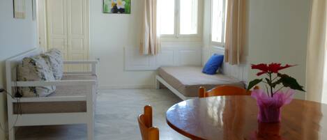A room with a wooden dining table, a white sofa, a window with curtains, and a small plant on the table.