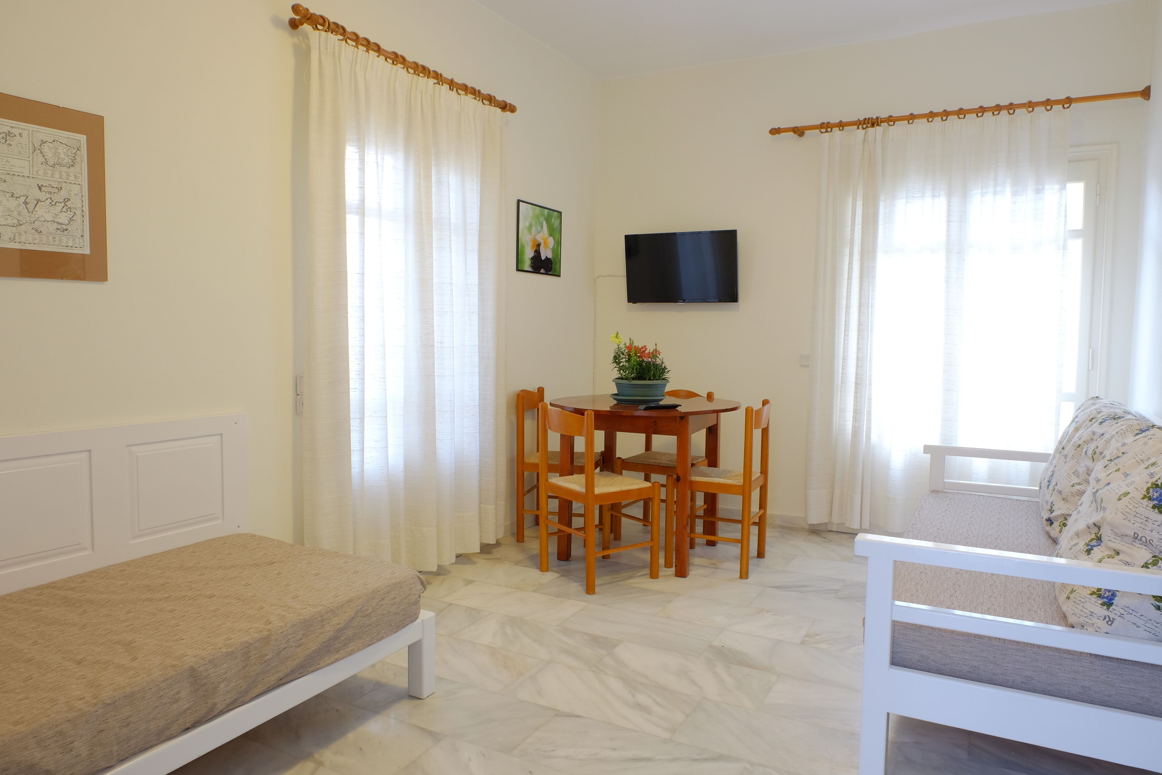 Una habitación pequeña y bien iluminada con cama, una mesa de comedor con sillas, televisor montado en la pared y ventana con cortinas.