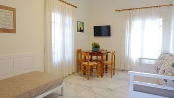 Una habitación pequeña y bien iluminada con cama, una mesa de comedor con sillas, televisor montado en la pared y ventana con cortinas.