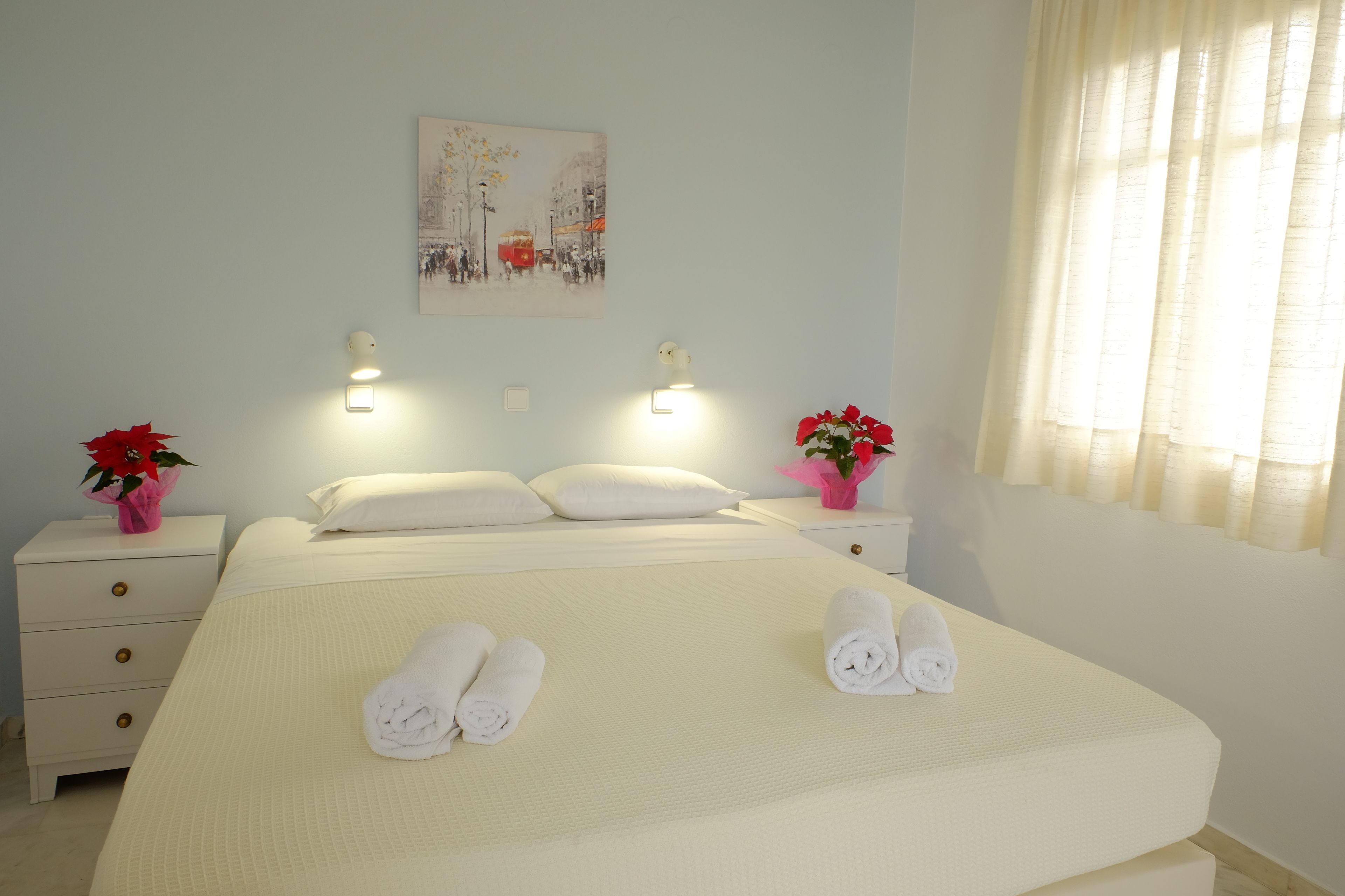 A neatly made bed with rolled towels, a painting on the wall, and two bedside tables with lamps and flowers.