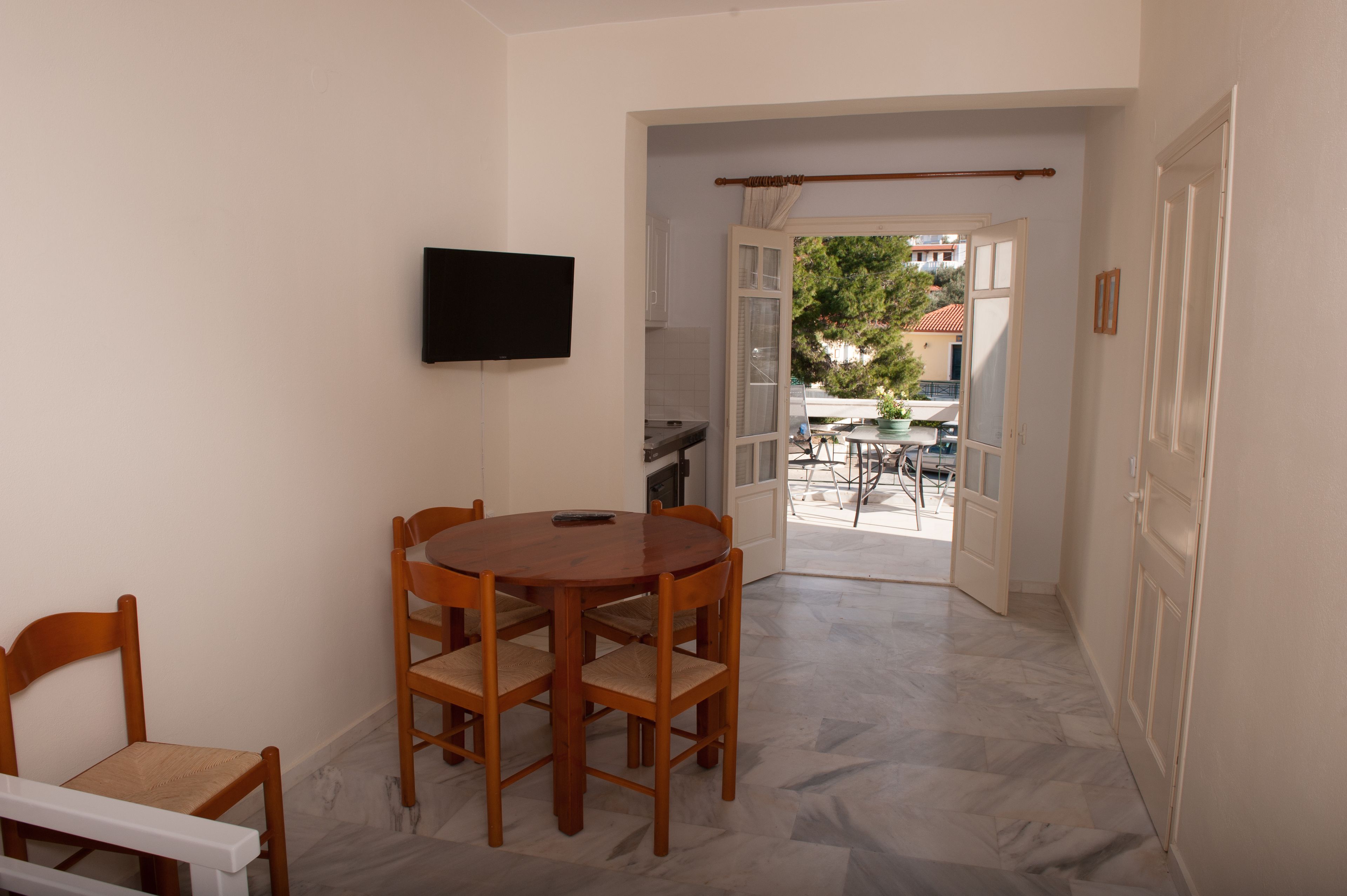 A room with a dining table and chairs, a TV mounted on the wall, and an open door leading to an outdoor area with a table and chairs.