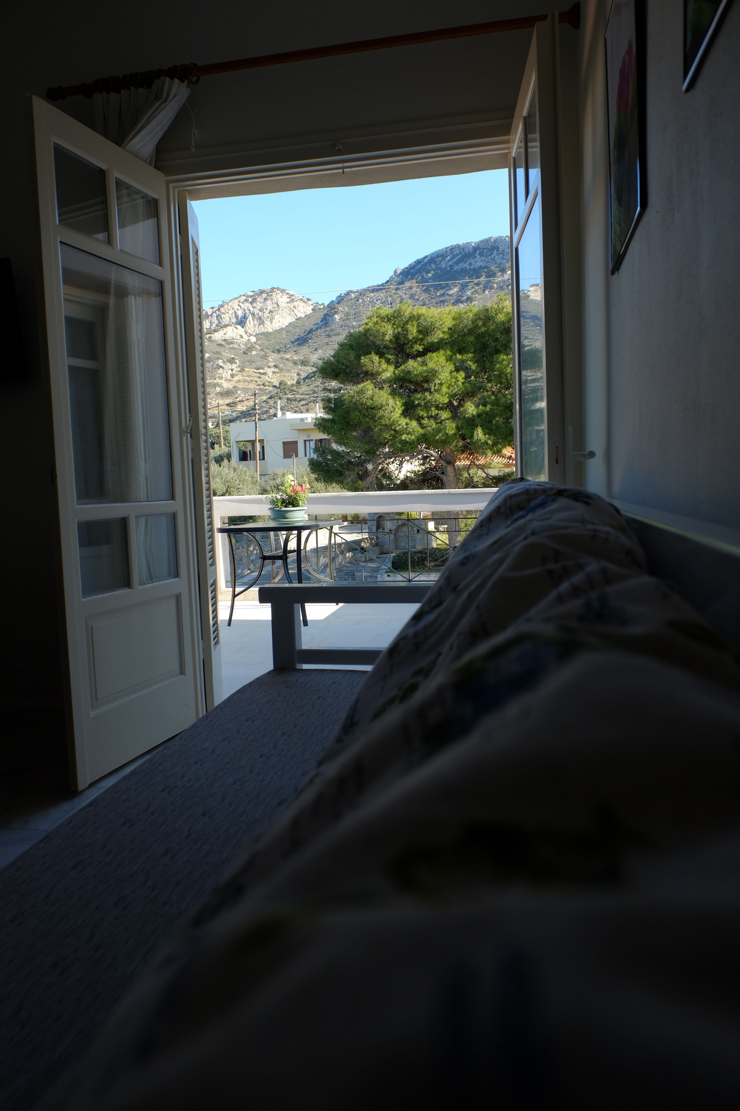 A room with a bed, a window, and a view of a balcony with a table and chairs.