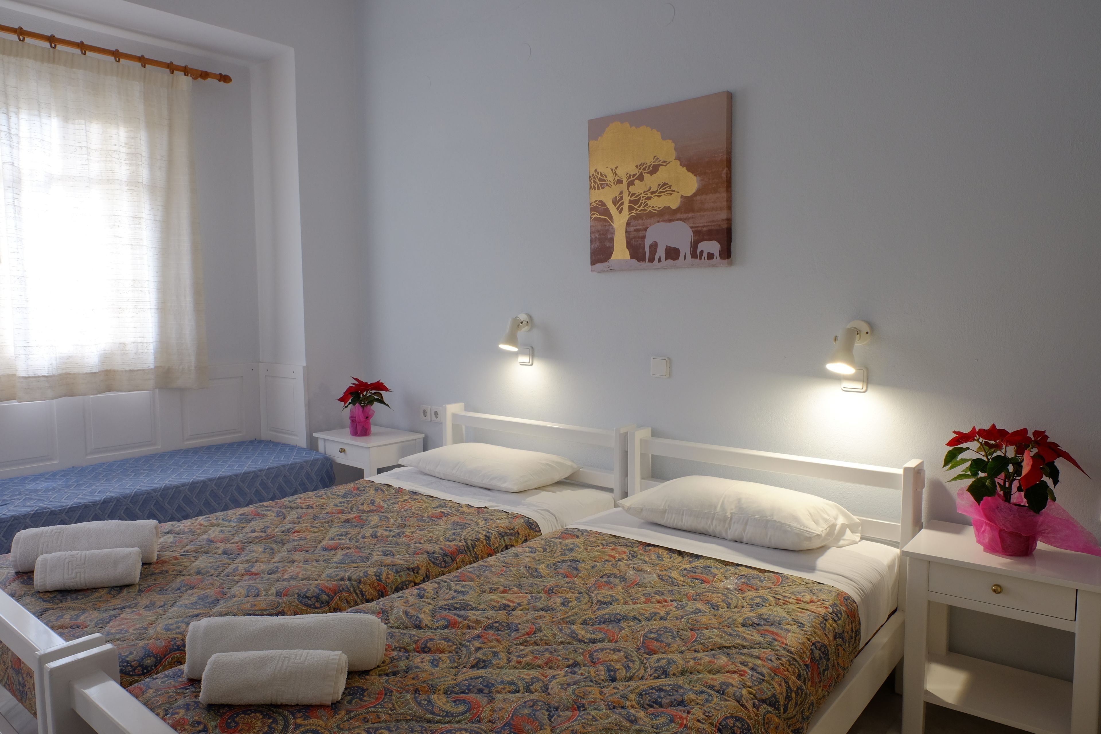 A bedroom with two beds, a window with curtains, a wall painting, bedside lamps, and a nightstand with a plant.