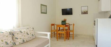 A small, bright room with a sofa, a dining table with chairs, a flat-screen TV, and a refrigerator.