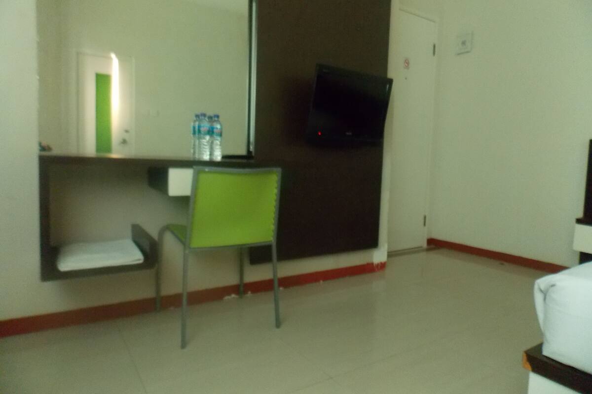A hotel room with a bed, a green chair, a TV mounted on the wall, and a water dispenser on the nightstand.
