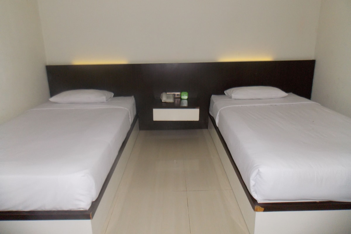 A hotel room with two single beds, a nightstand with a phone and a green clock, and a lighted wall above the beds.