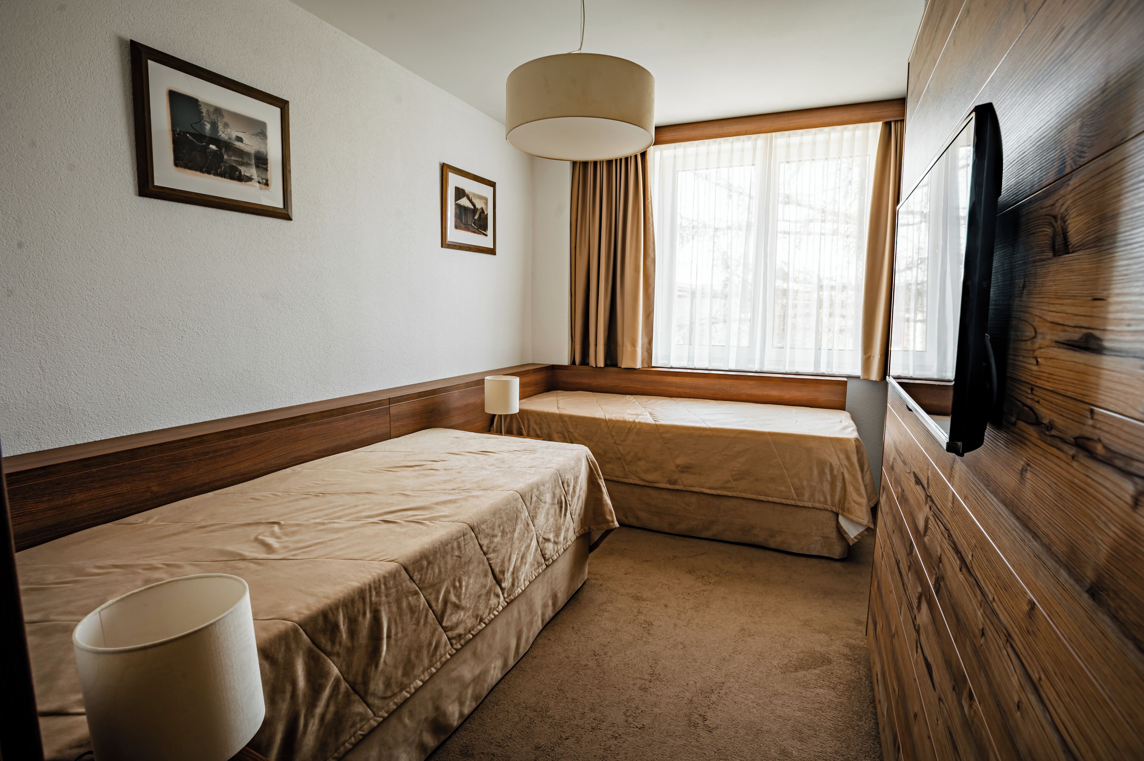 A hotel room with two single beds, a window with curtains, a mounted TV, and framed pictures on the wall.