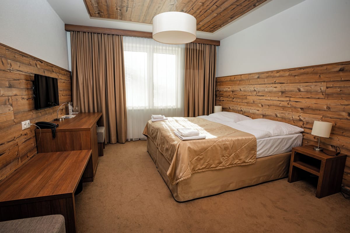 Hotelkamer met houten hoofdeinde, bed met beige sprei, nachtkastje met lamp, bureau met flatscreen televisie en groot raam met gordijnen.