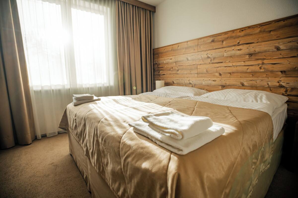 Een hotelkamer met een houten hoofdeinde, een groot bed met beige beddengoed en een raam met transparante gordijnen.
