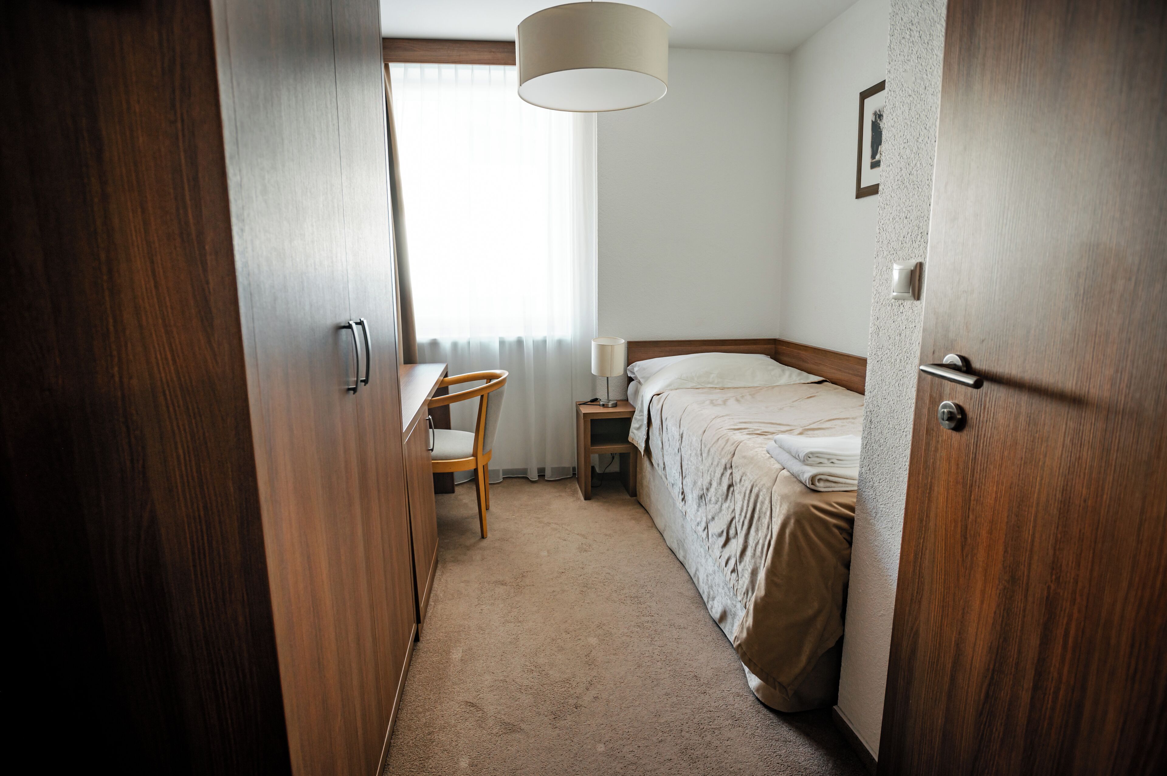 A hotel room with a wooden wardrobe, a bedside table with a lamp, a chair, and a bed with a beige blanket.