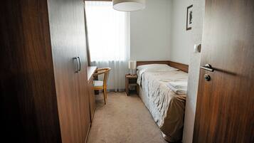 A hotel room with a wooden wardrobe, a bedside table with a lamp, a chair, and a bed with a beige blanket.