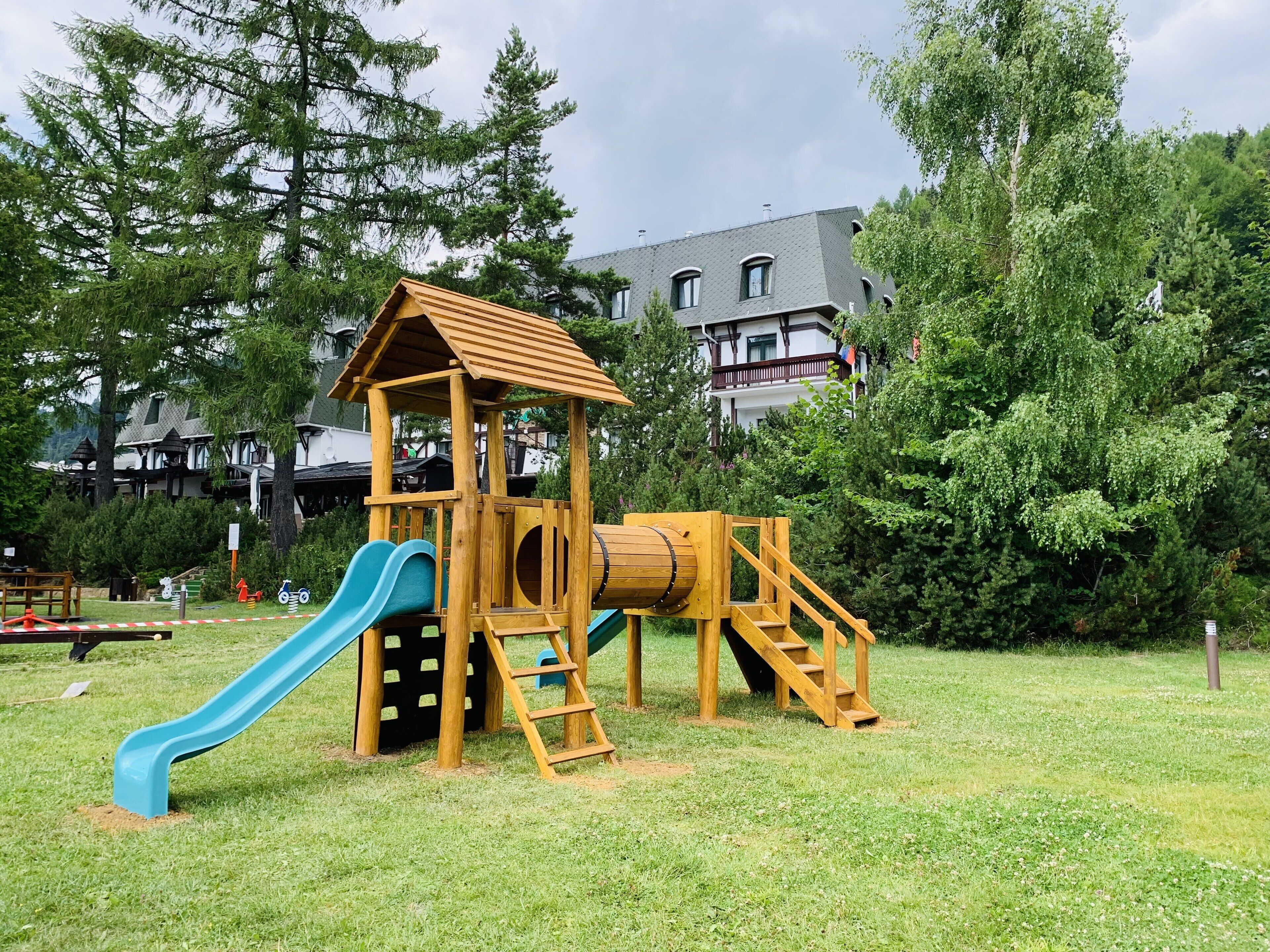 Une structure de jeux en bois, dotée d’un toboggan et d’éléments d’escalade, entourée de pelouse et d’arbres, avec un bâtiment en arrière-plan.
