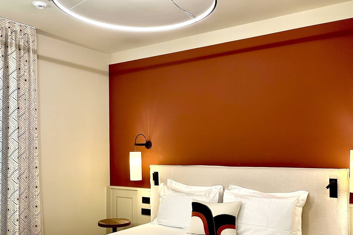 Une chambre d’hôtel moderne avec un grand lit, un luminaire rond au plafond, une table de chevet et une lampe fixée au mur.