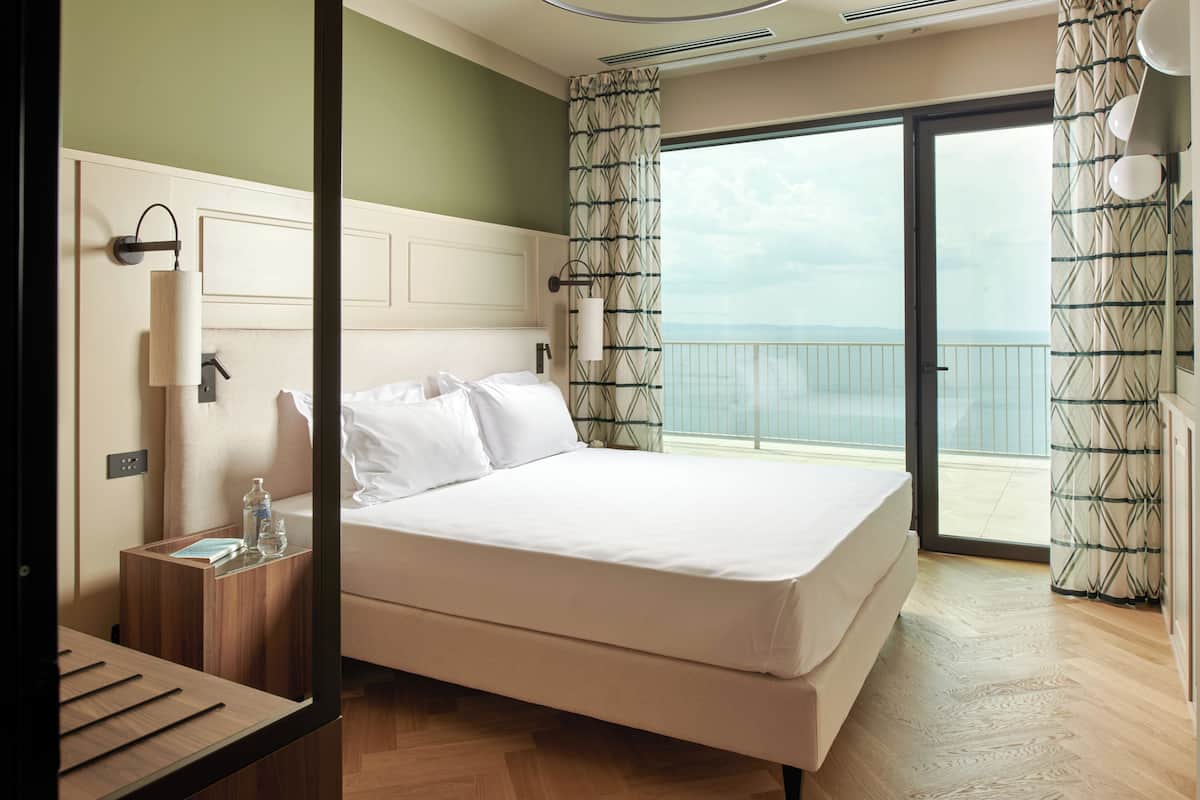 Une chambre d’hôtel avec un lit, une table de chevet et une vue sur la mer, visible à travers une grande fenêtre.