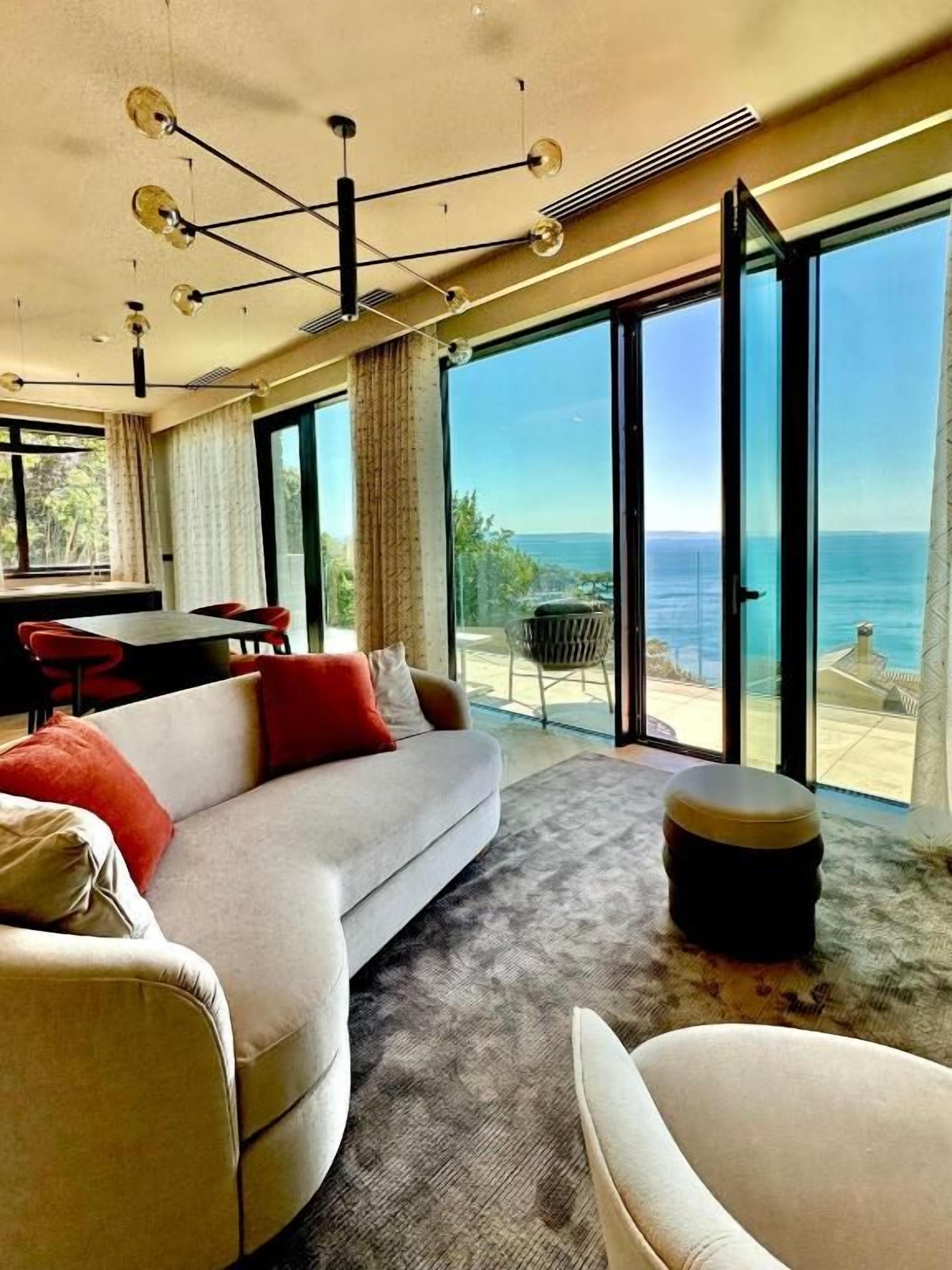 Ein modernes Wohnzimmer mit Sofa, Hocker und Essbereich, große Fenster mit Meerblick.