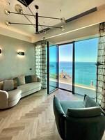 Un soggiorno moderno con pavimento in legno a spina di pesce, un divano, una poltrona verde e un'ampia porta scorrevole in vetro che dà su un balcone con vista sul mare.