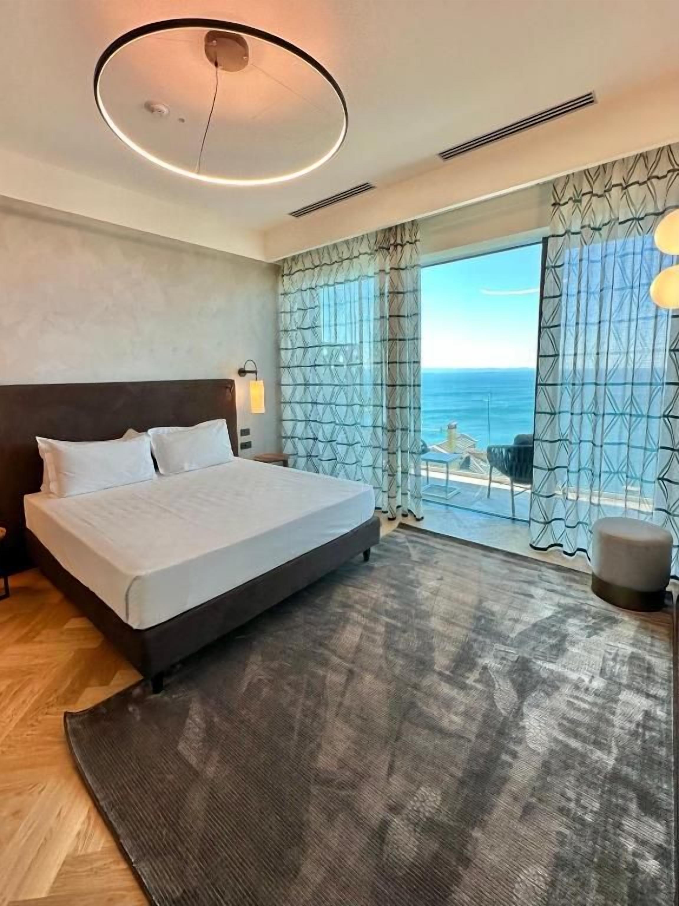 Habitación de hotel con cama, vista al mar y un ventanal grande con cortinas.