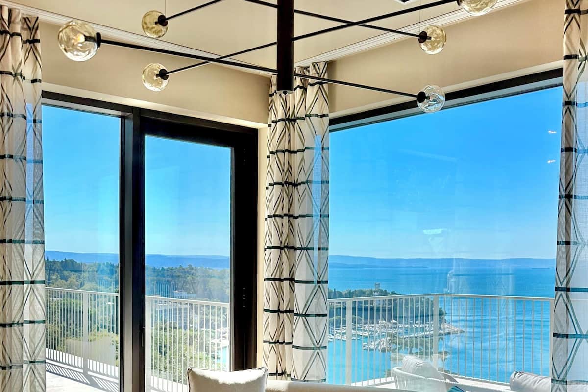 Un salon moderne avec un canapé beige, un pouf rond et une petite table. De grandes fenêtres offrent une vue sur la mer et sur le paysage urbain au loin.