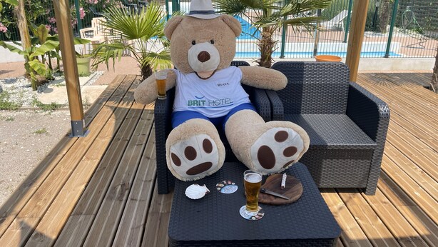 A large teddy bear seated on a wicker lounge chair with a glass of beer and a coaster on a wooden deck.