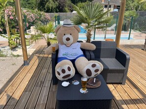 A large teddy bear seated on a wicker lounge chair with a glass of beer and a coaster on a wooden deck. - Brit Hotel Brive (Malemort-sur-Correze)