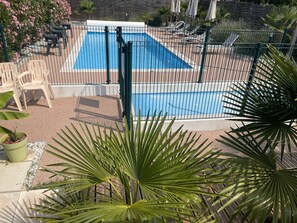 Une piscine rectangulaire entourée d’une clôture noire, bordée d’une terrasse aménagée avec des chaises et des plantes en pot.