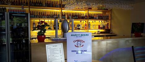 Zona de bar con una variedad de botellas y vasos en estanterías. Un cartel con la carta es visible en primer plano.