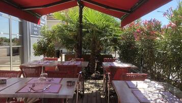 Un área de comedor al aire libre con mesas y sillas, una palmera en el centro y un paraguas rojo.