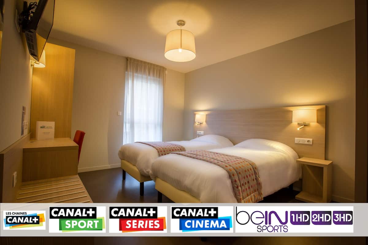 Une chambre d’hôtel avec deux lits, un bureau et une chaise. Elle est équipée de lampes fixées au mur et d’un luminaire au plafond.