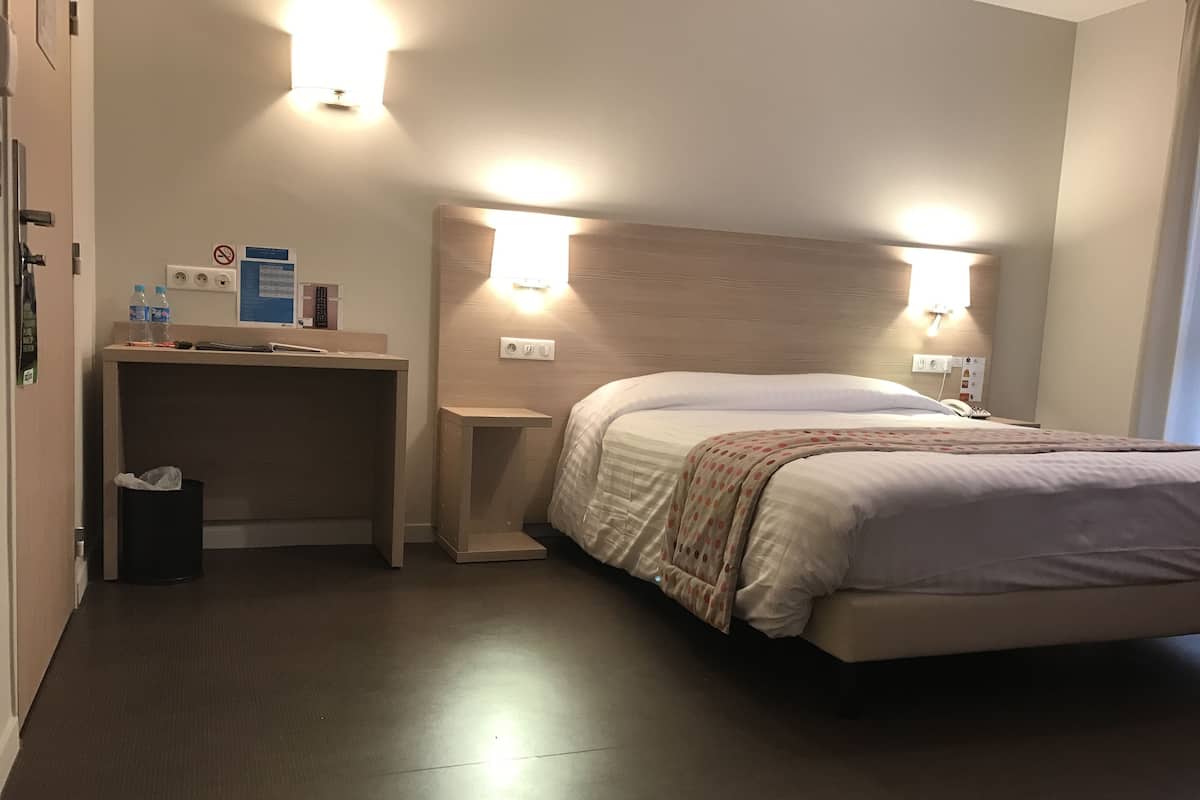 Une chambre d’hôtel avec un lit, des tables de chevet, un petit bureau et une poubelle.