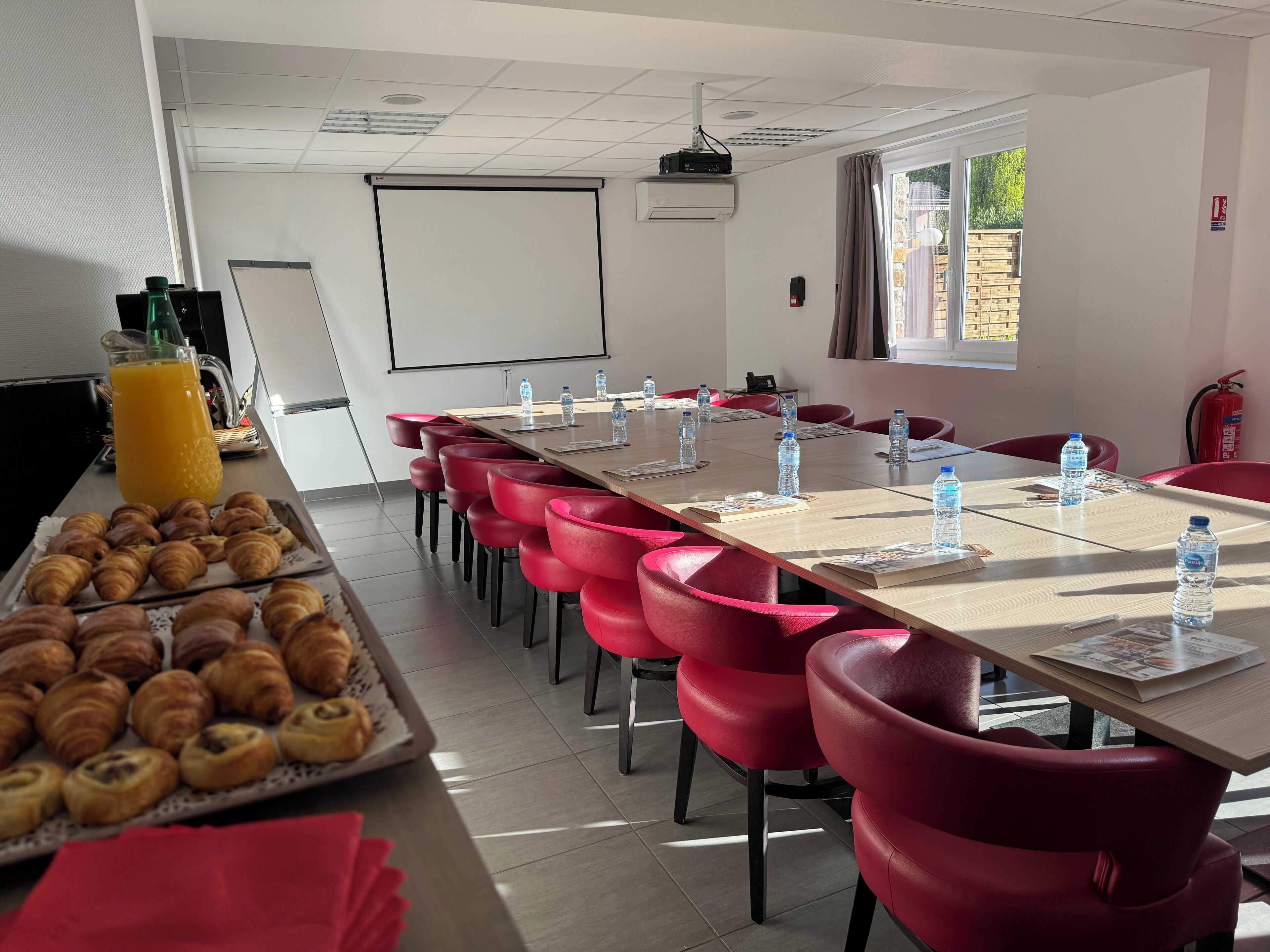 Ein Konferenzraum mit einem langen Tisch, roten Stühlen, Wasserflaschen und einer Auswahl an Gebäck.