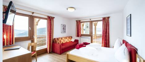 A hotel room with a bed, a desk, a chair, a sofa, and large windows with curtains.