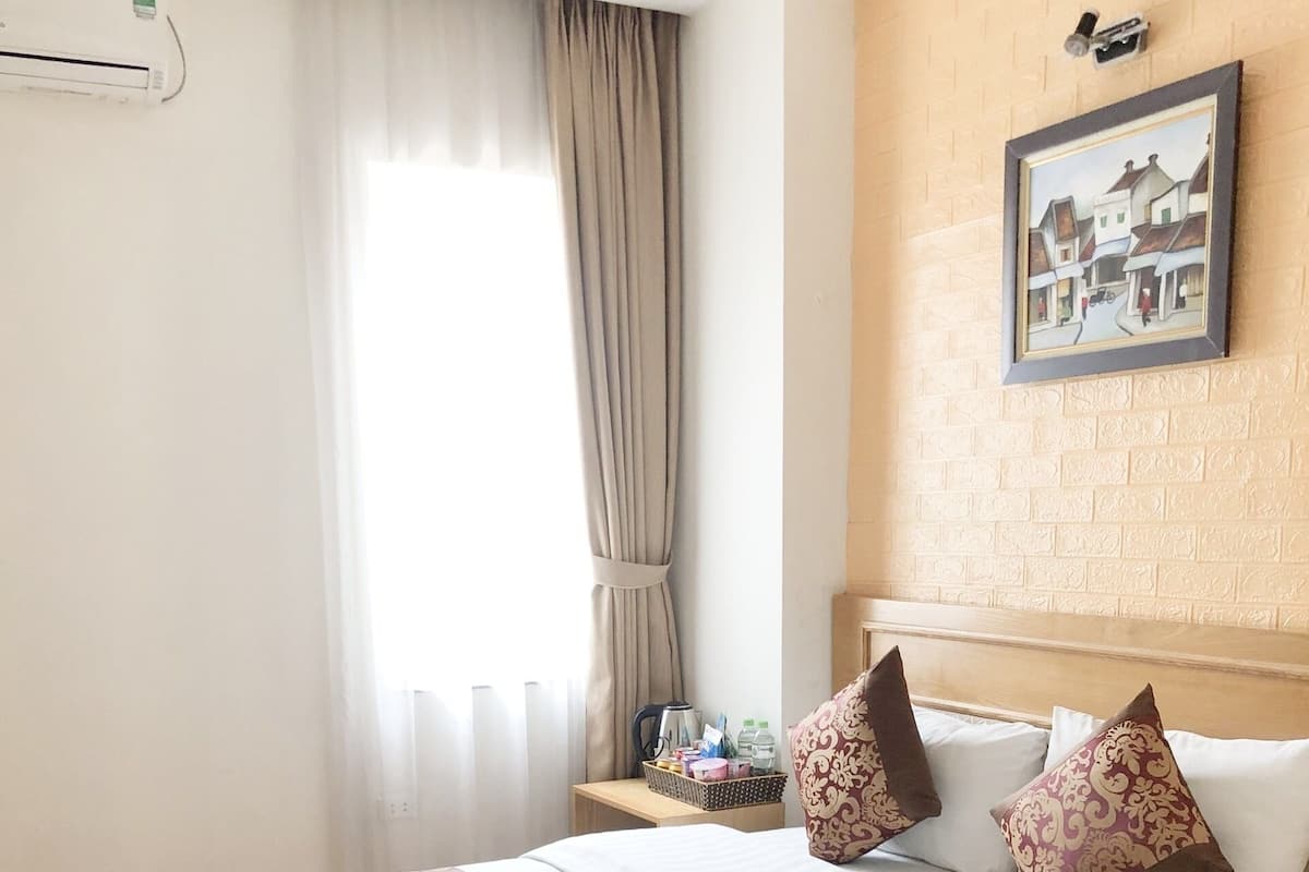 A hotel room with a large bed, a headboard with a painting, a bedside table with toiletries, and a window with curtains.
