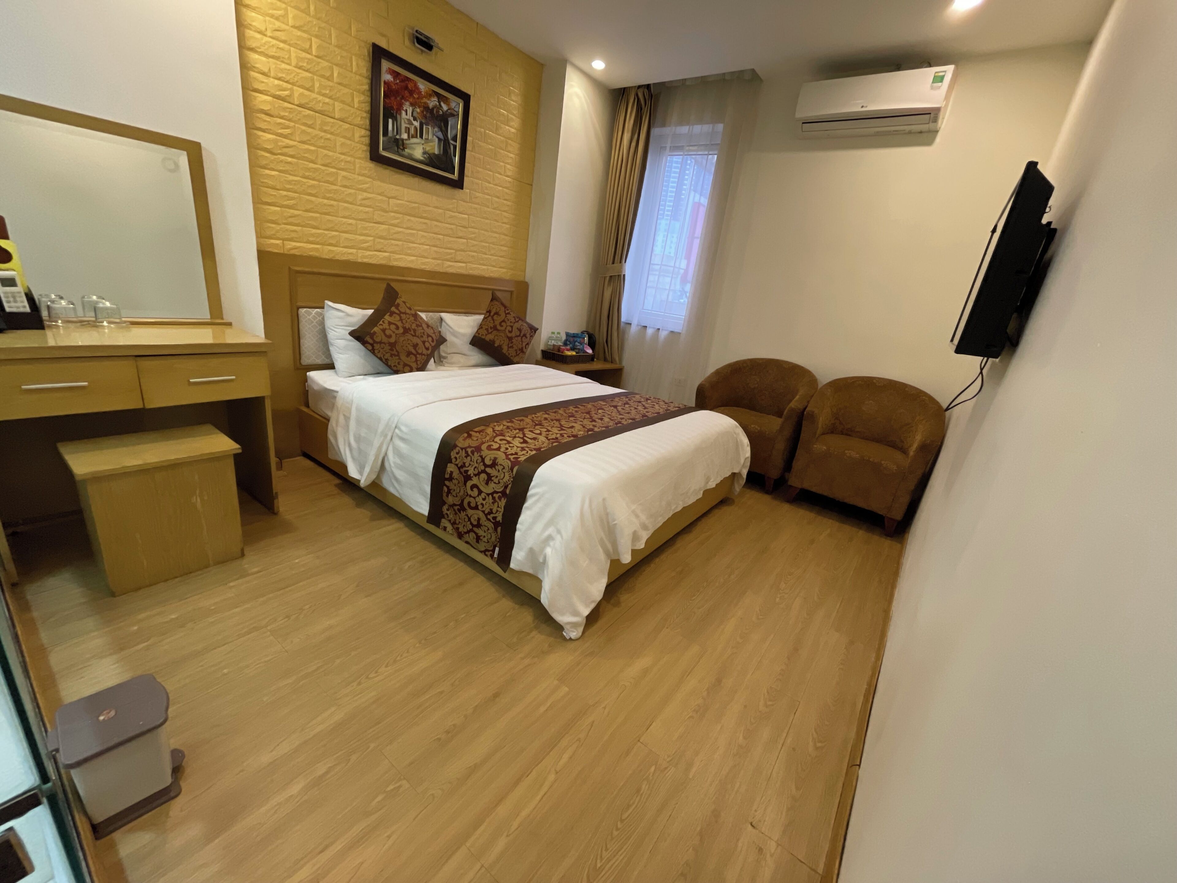 A hotel room with a bed, a desk, two armchairs, a small table, a mirror, a painting on the wall, and an air conditioning unit.