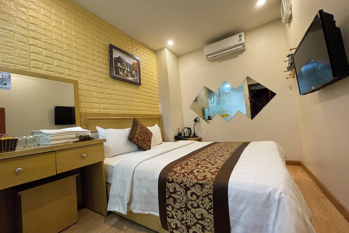 A hotel room with a bed, a desk with a mirror, a wall-mounted air conditioner, and a TV.