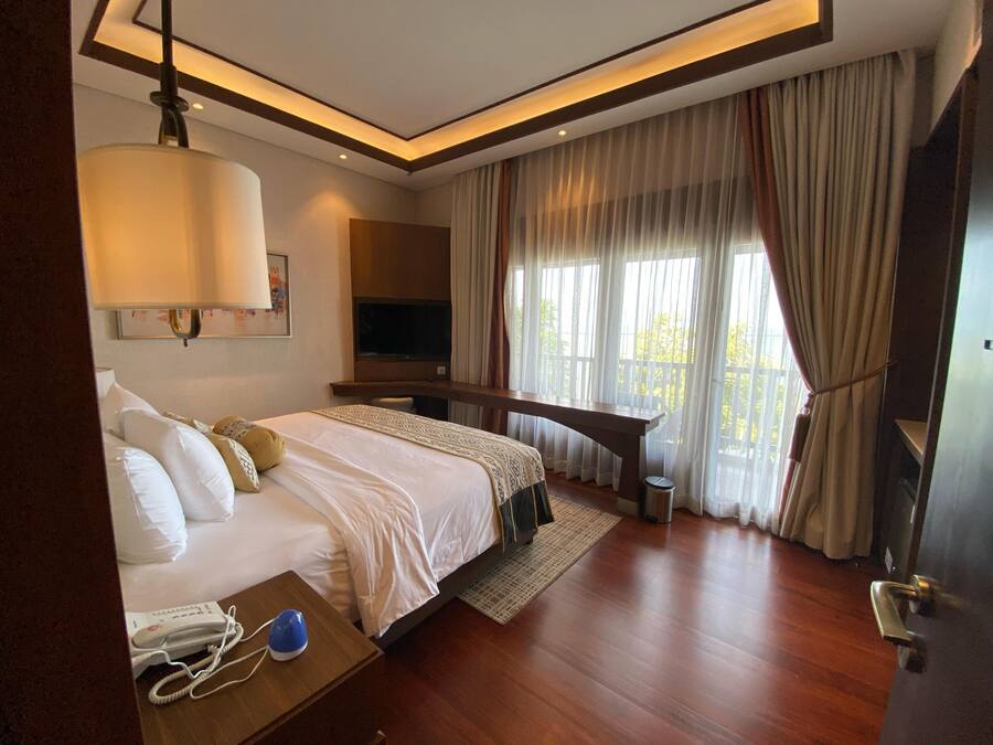 A hotel room with a bed, bedside table, lamp, TV, and large windows with curtains.